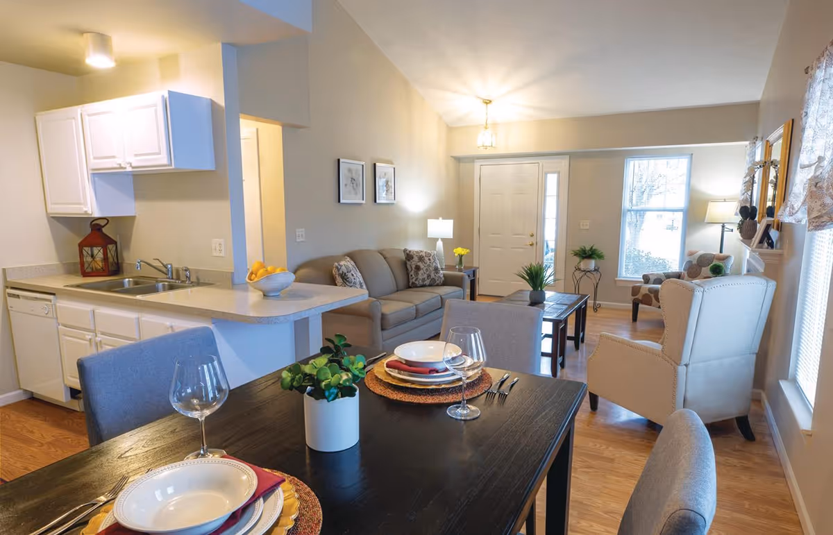 A bright and cozy living space featuring a small kitchen with white cabinets and a countertop, a dining table set with plates, glasses, and a small plant centerpiece, and a living room area with a sofa, armchair, coffee table, and decorative plants. The room has wooden flooring, neutral-colored walls, and windows allowing natural light to enter.