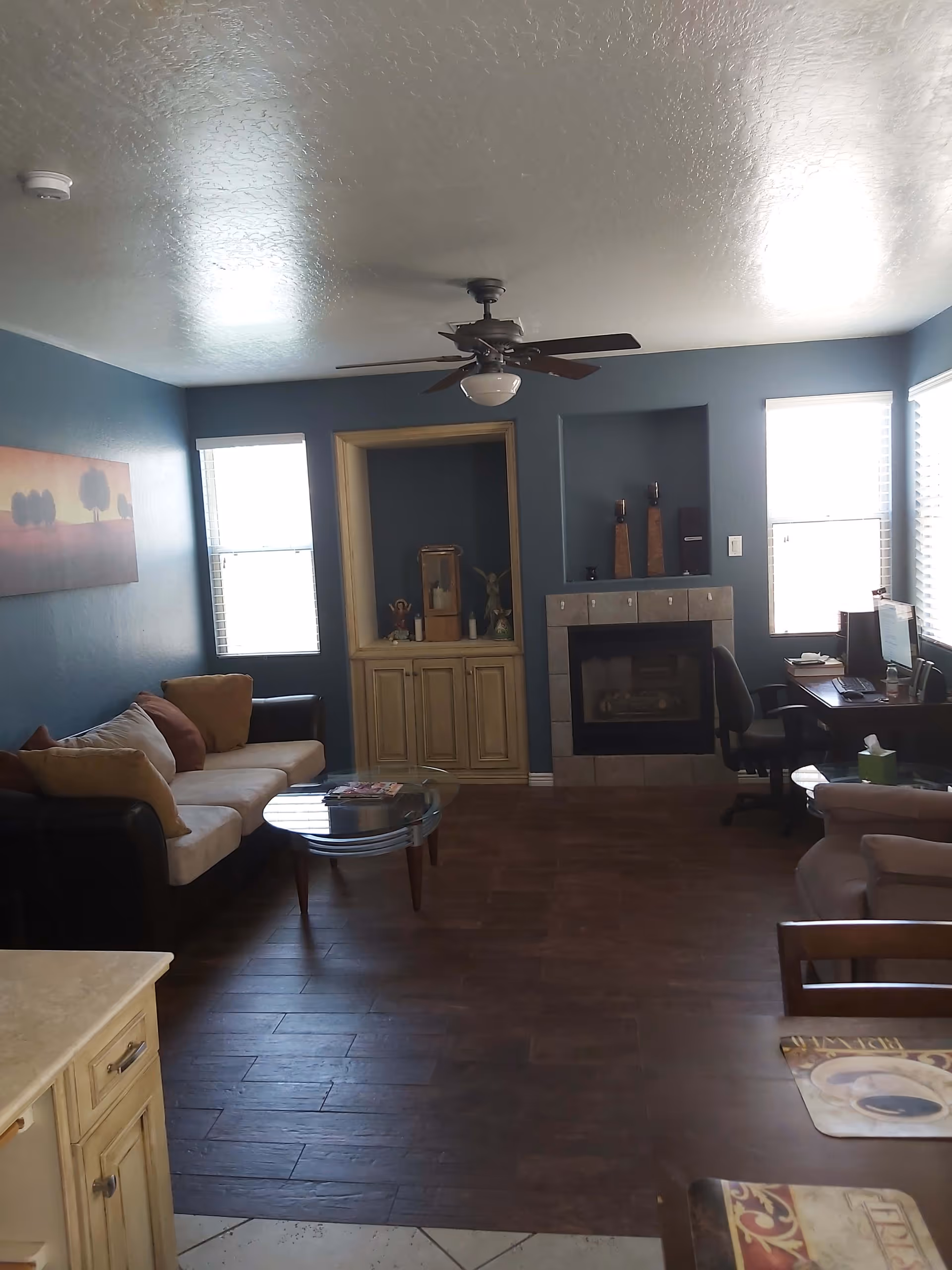 A living room with blue walls, two windows with blinds, a ceiling fan, a beige and brown sofa with cushions, a glass coffee table, a fireplace with decorative items on the mantel, a computer desk with a chair and computer, and a beige armchair partially visible. There is also a painting of trees on the left wall.