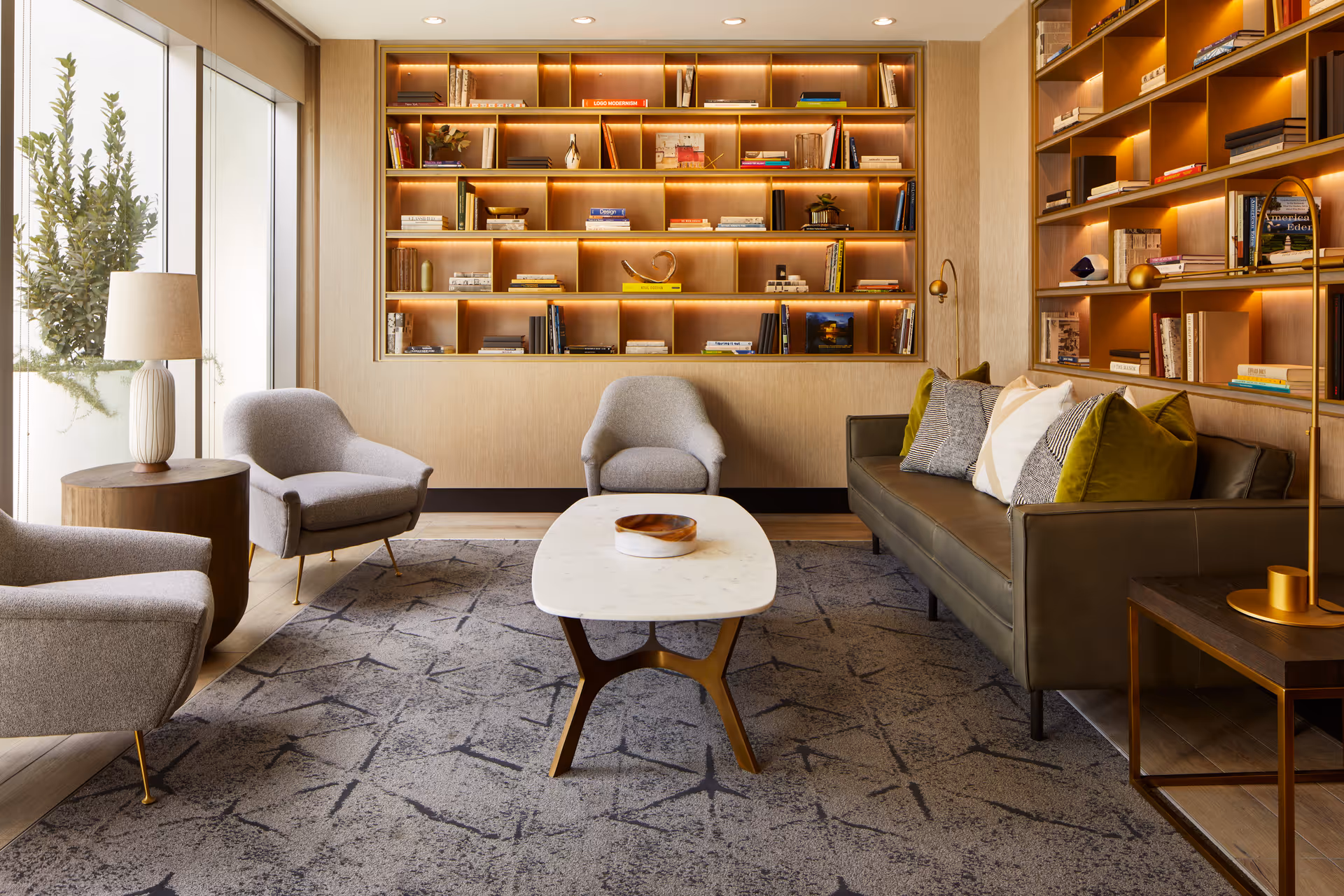 Sunlit lounge with a sofa, armchairs, a coffee table, and illuminated built-in bookshelves beside a large window.