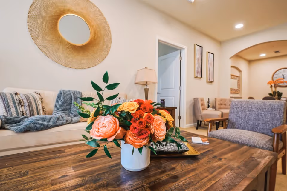A cozy living room with a wooden coffee table displaying a vase of orange and pink roses, with sofas and chairs in the background.