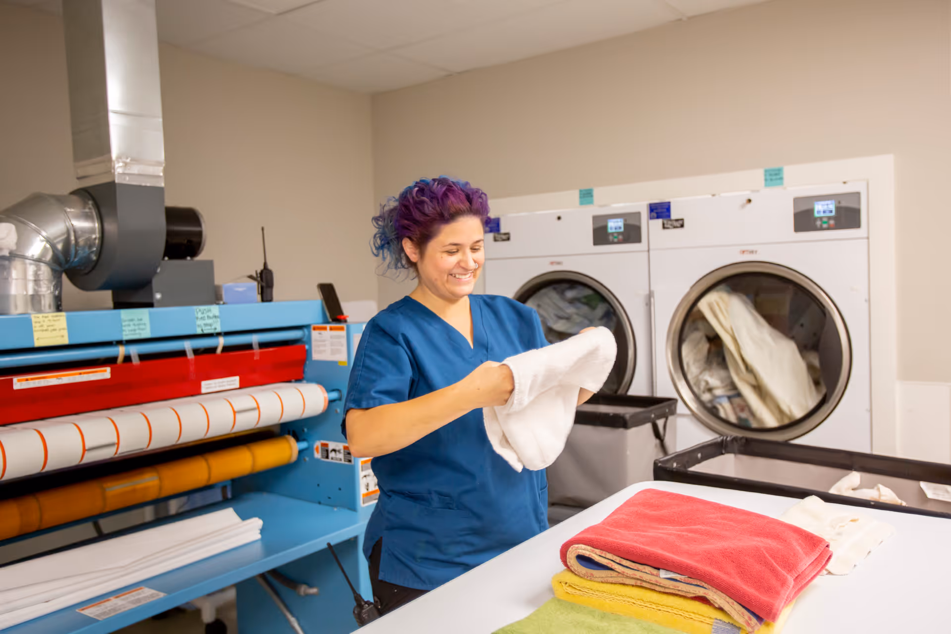 A woman with purple and blue hair wearing blue scrubs is folding a white towel in a laundry room. Behind her are two large front-loading dryers filled with laundry, and to the left is a large blue ironing or folding machine. On the table in front of her are neatly folded towels in red, yellow, and green.