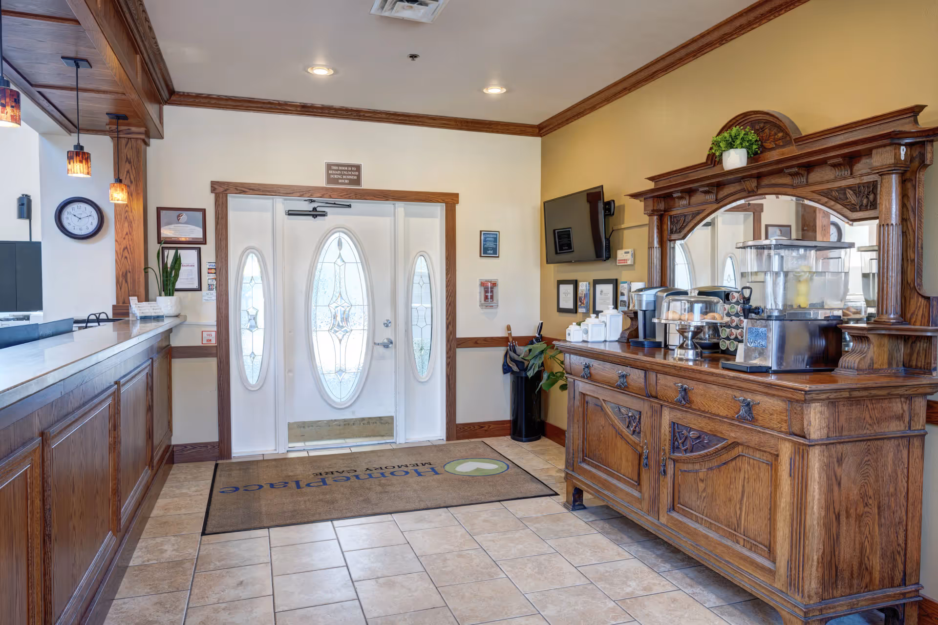 Interior view of a senior living facility entrance area featuring a wooden reception desk on the left and a large ornate wooden sideboard with a mirror on the right. The sideboard holds a water dispenser, coffee machine, and a tray of pastries. The entrance door has decorative glass panels and a mat on the floor with the text 'HomePlace Memory Care'. The walls are light-colored with wooden trim, and there is a wall-mounted TV and framed certificates on the right wall.