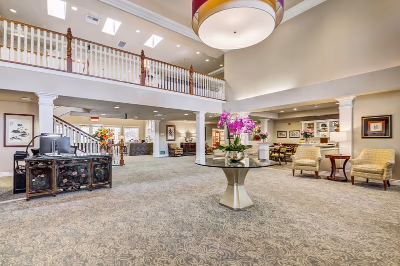 Spacious and well-lit senior living facility common area with high ceilings, a large round table with a vase of purple orchids in the center, comfortable seating including armchairs and sofas, decorative columns, a staircase leading to an upper level with a white railing, and various framed artworks on the walls.