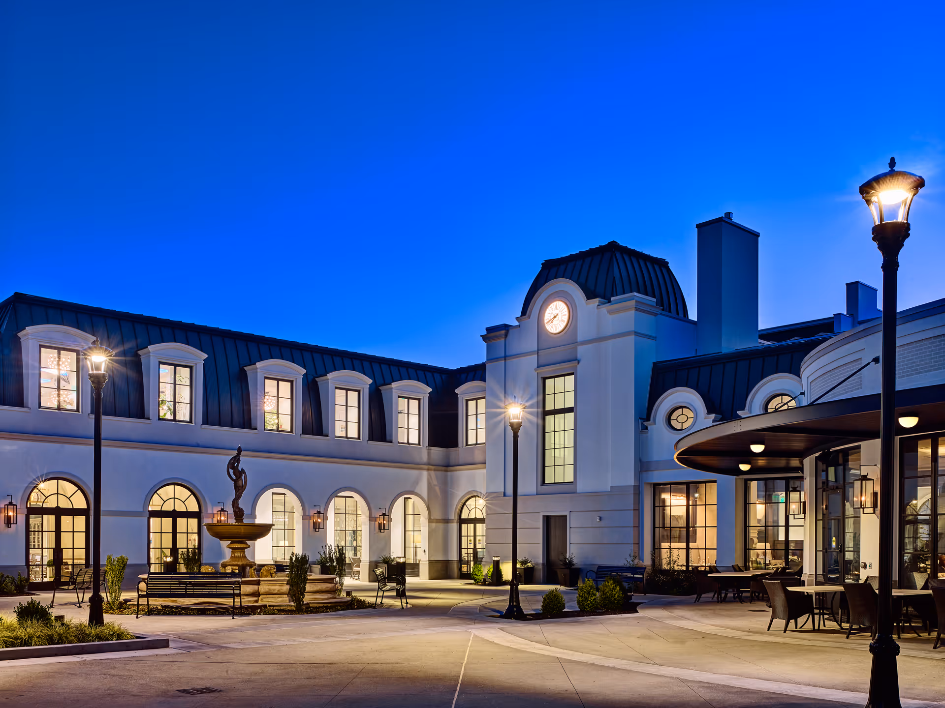 Exterior view of a senior living facility at dusk with a courtyard featuring a fountain, benches, and outdoor seating. The building has large arched windows, a clock tower, and street lamps illuminating the area.