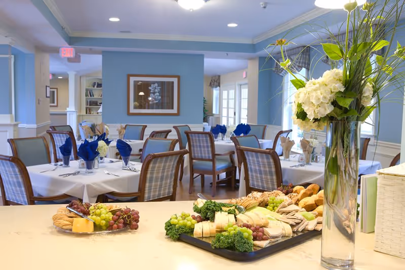 A bright and elegant dining room with tables set with white tablecloths, blue and beige napkins, and wooden chairs with plaid upholstery. In the foreground, there is a countertop with trays of assorted bread, cheese, grapes, and greenery, along with a tall glass vase holding a bouquet of white flowers and green foliage.