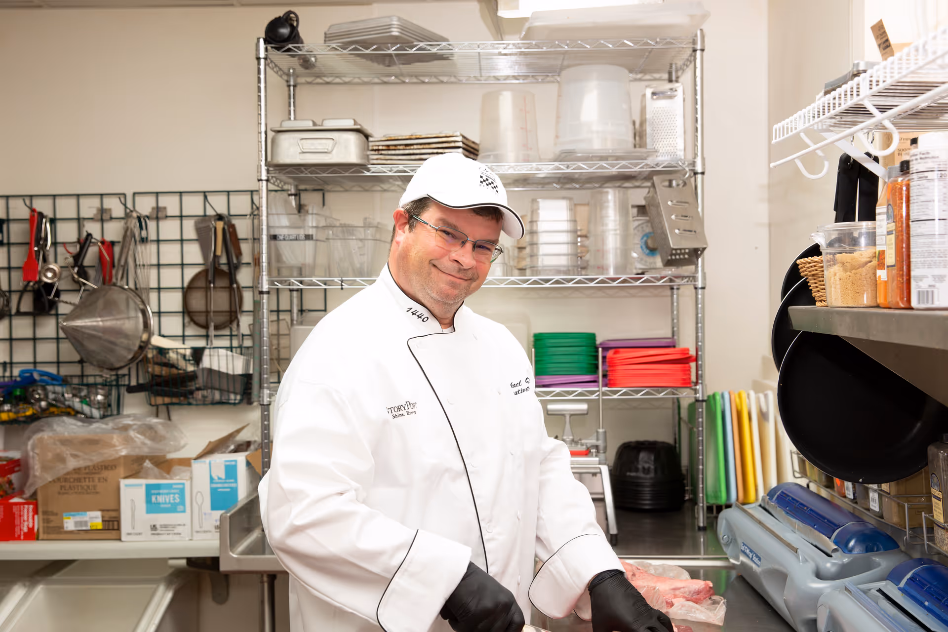 A chef wearing a white uniform, white cap, and black gloves is standing in a kitchen area with metal shelves holding various kitchen utensils, containers, and cutting boards. The chef is smiling and preparing food on a counter.
