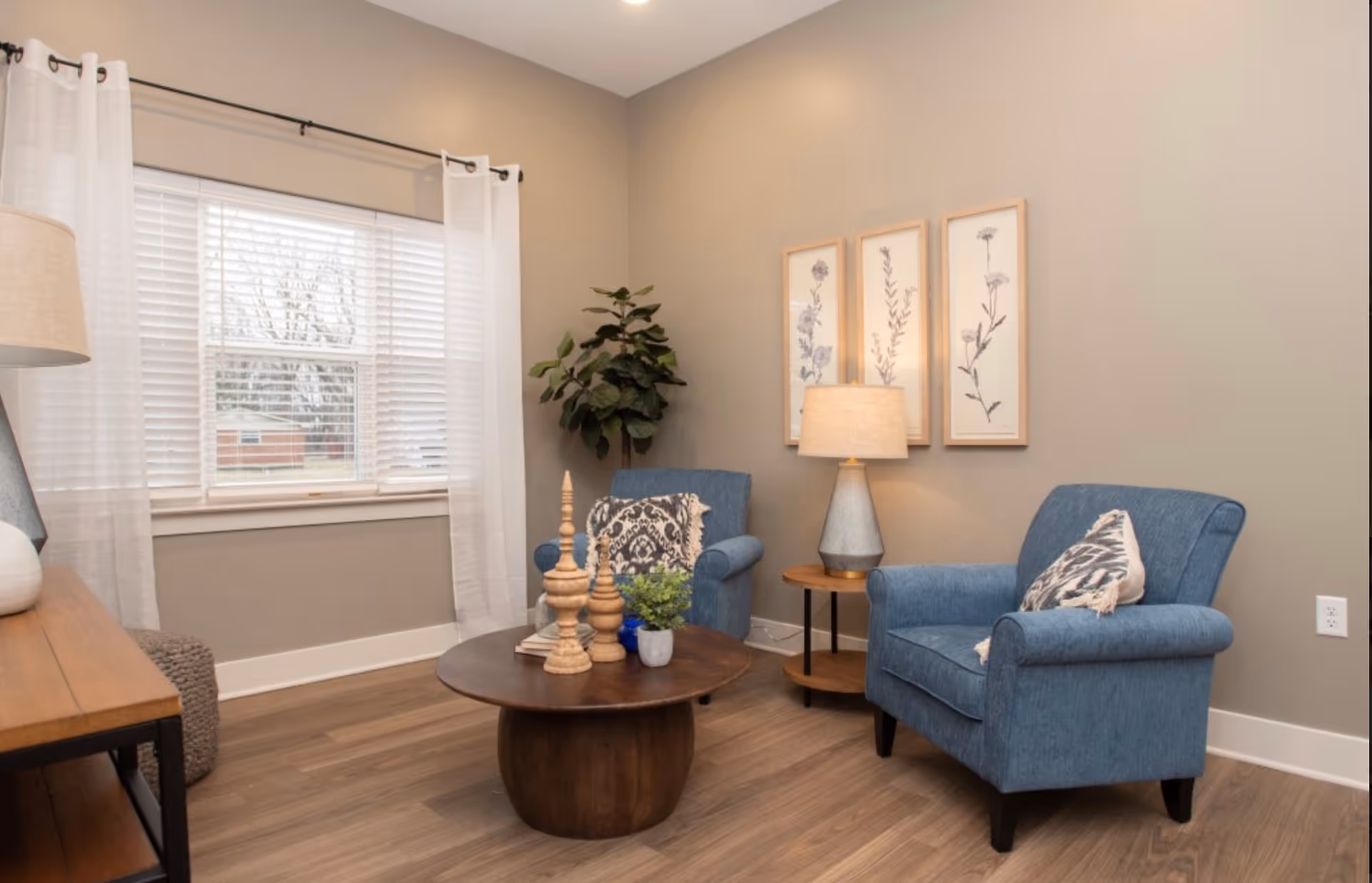 A cozy living room area with two blue upholstered armchairs, each with a decorative pillow. Between the chairs is a round wooden side table with a lamp. In front of the chairs is a round wooden coffee table with decorative items and a small plant. The room has light brown walls, wood flooring, a large window with white blinds and sheer white curtains, and three framed botanical prints on the wall. A tall green plant is placed in the corner near the window.