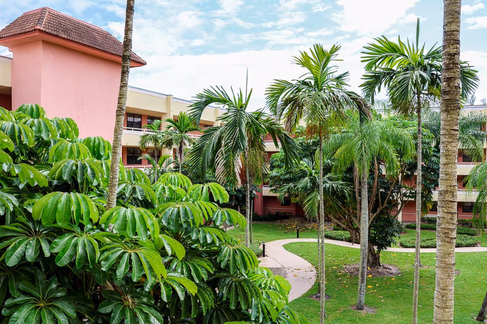 Outdoor view of a senior living facility courtyard with lush green tropical plants and palm trees, a winding paved walkway, and a multi-story building with pink and beige walls in the background under a partly cloudy sky.