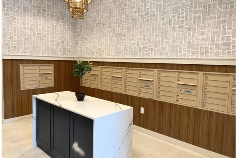 Interior view of a mailroom with multiple beige mailboxes mounted on wood-paneled walls. A white marble-topped island with black cabinetry stands in the center, decorated with a small potted plant. The upper walls have a textured wallpaper with a pattern resembling newspaper text, and a gold chandelier hangs from the ceiling.
