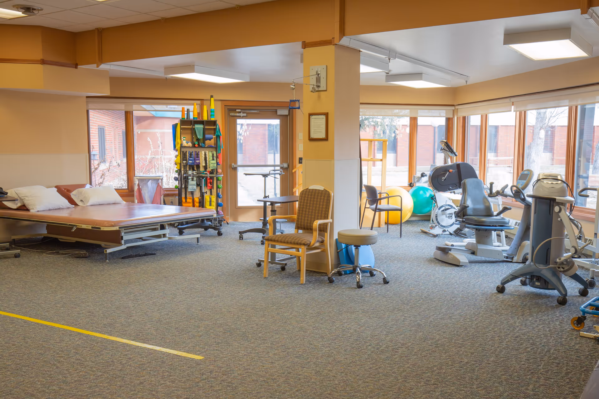 A spacious rehabilitation or physical therapy room with exercise equipment including stationary bikes, therapy balls, and a large adjustable therapy bed. The room has large windows allowing natural light, a few chairs, and a rack with various therapy tools and supplies.