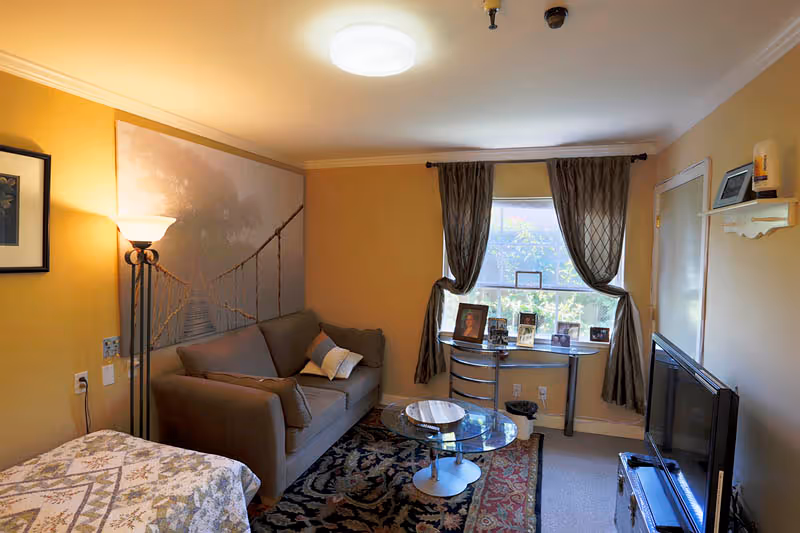 A cozy living room with a brown sofa, a glass coffee table on a patterned rug, a floor lamp, and a window with dark curtains. There is a small table under the window with framed photos and a TV on a stand to the right. The walls are painted a warm yellow color.