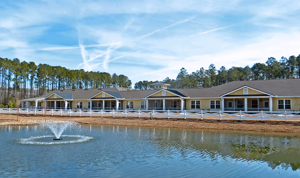 Exterior view of a single-story senior living facility building with a white fence in front and a pond with a water fountain in the foreground. The building is surrounded by trees under a blue sky with some clouds.