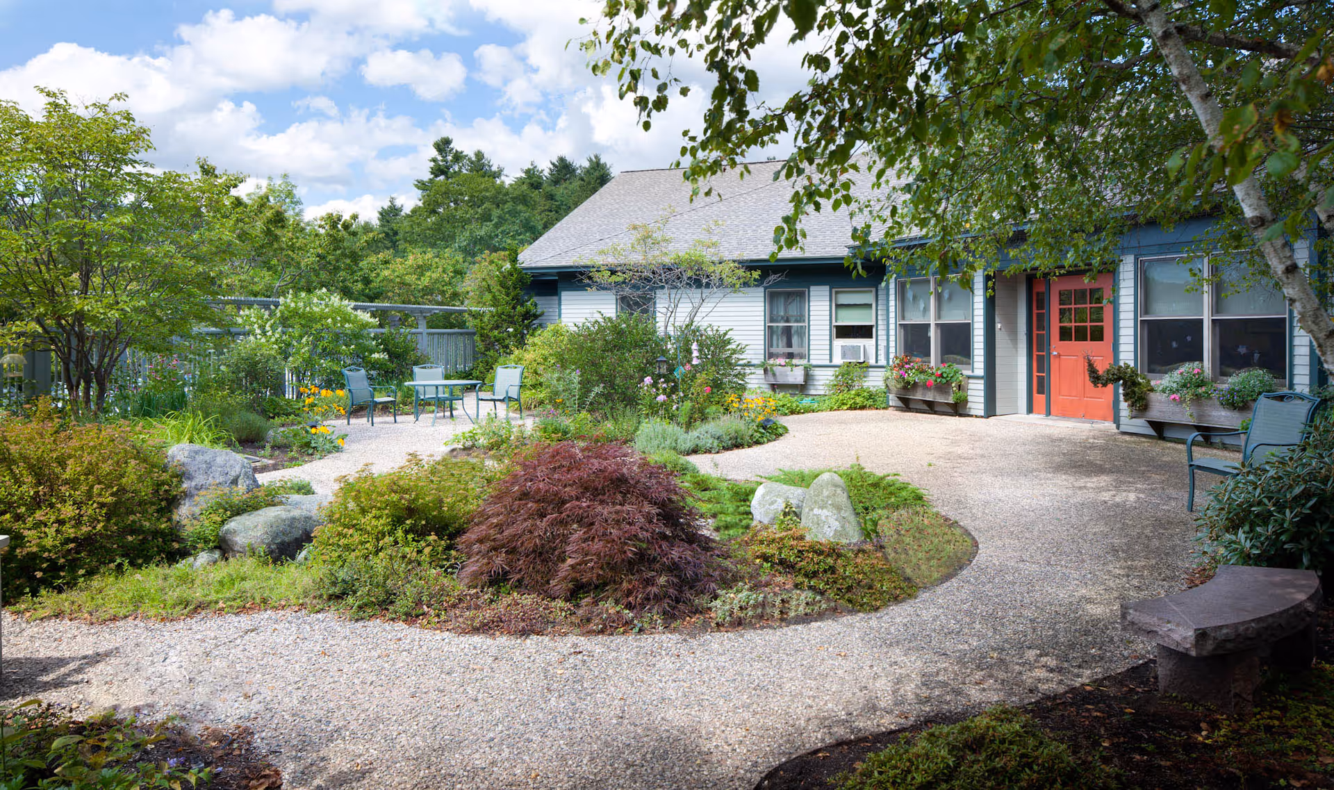 A landscaped courtyard with gravel paths, garden beds, outdoor seating, and the front entrance of a single-story building with a red door.