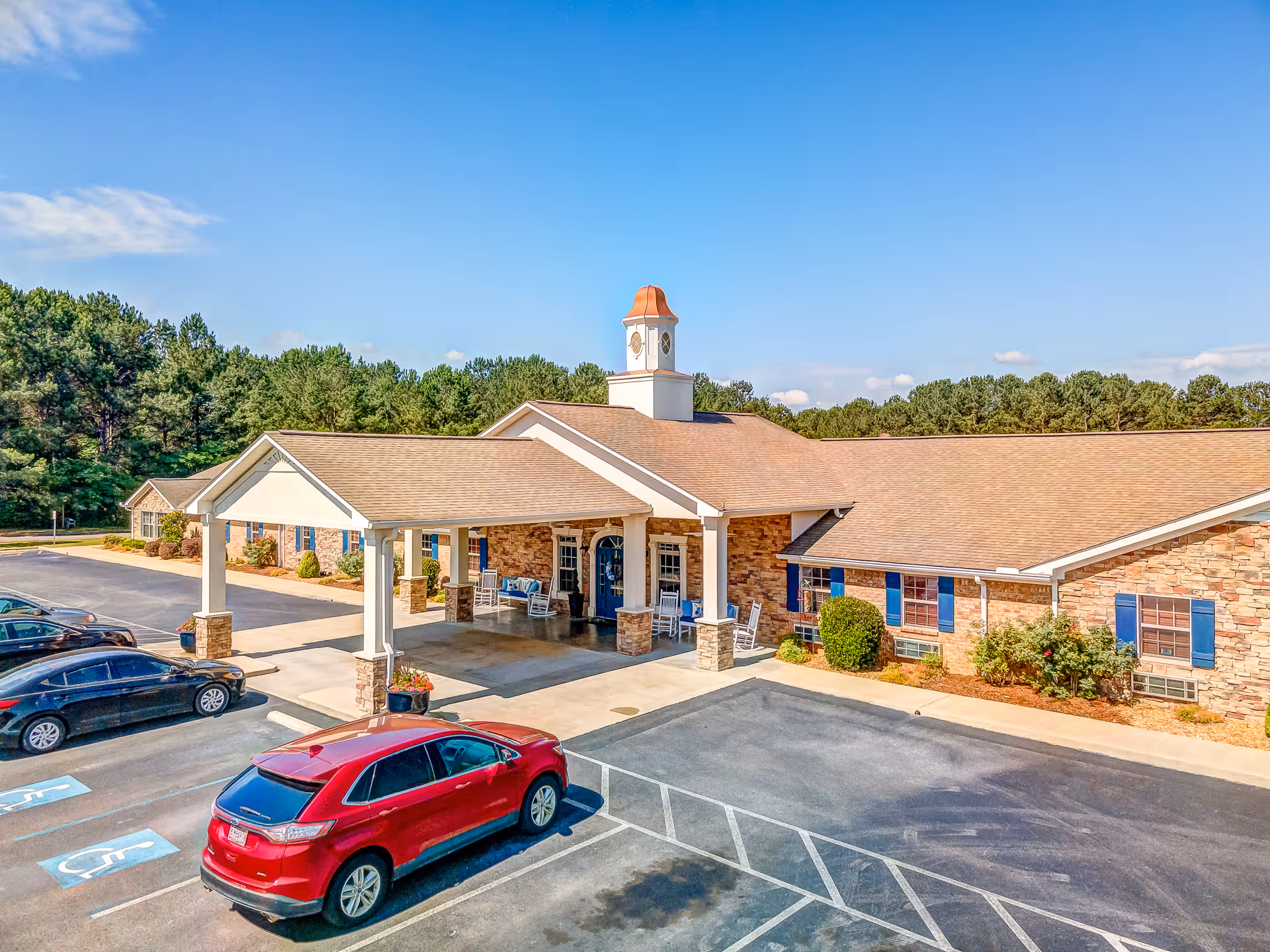Exterior view of Legacy Village of Jacksonville showing a single-story building with a covered entrance supported by columns. The building has a stone facade with blue window shutters and a small cupola on the roof. Several cars are parked in the parking lot in front of the building, including a red SUV in a handicapped parking space. Trees and a clear blue sky are visible in the background.