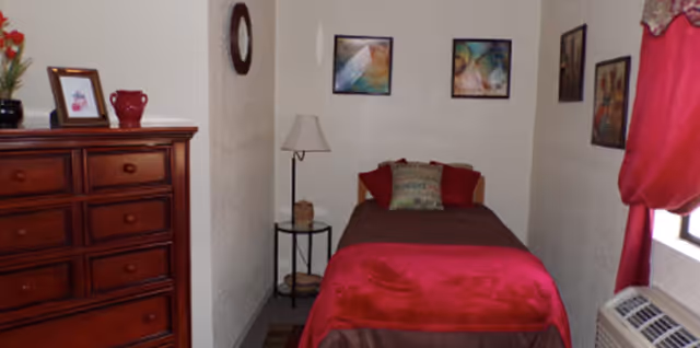 Small single-occupancy bedroom with a single bed dressed in red and brown linens, a wooden dresser, side table with lamp, and framed wall art.