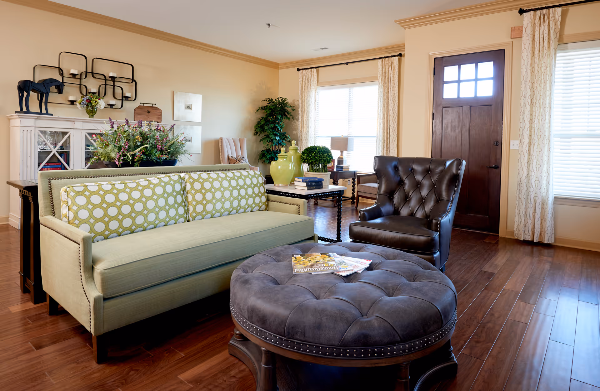 A cozy living room with a green sofa featuring patterned cushions, a dark brown leather armchair, and a large round tufted ottoman with magazines on top. The room has wooden flooring, a wooden front door with window panes, large windows with light curtains, and decorative plants and artwork on the walls.