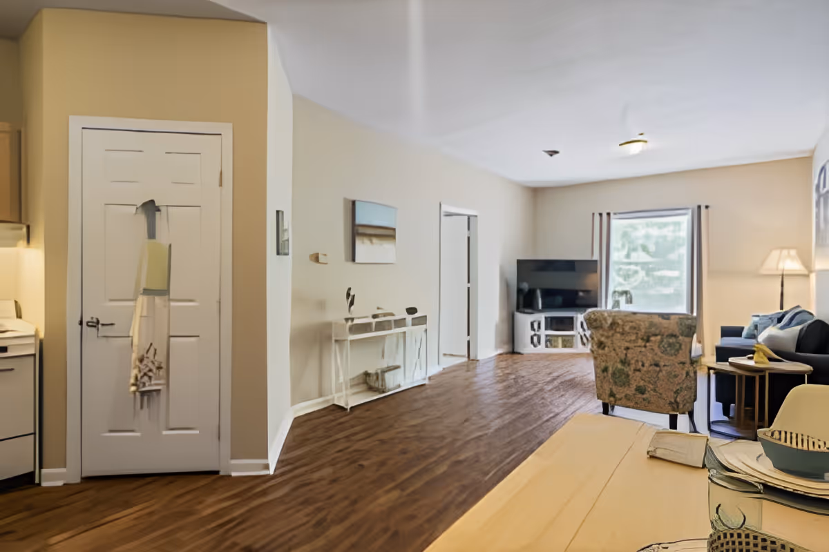 A spacious living room area with wood flooring, beige walls, and a large window with curtains. The room features a floral armchair, a dark blue sofa with cushions, a floor lamp, a TV on a white stand, and a small table with a decorative bowl and book. To the left, part of a kitchen and a closed white door are visible.
