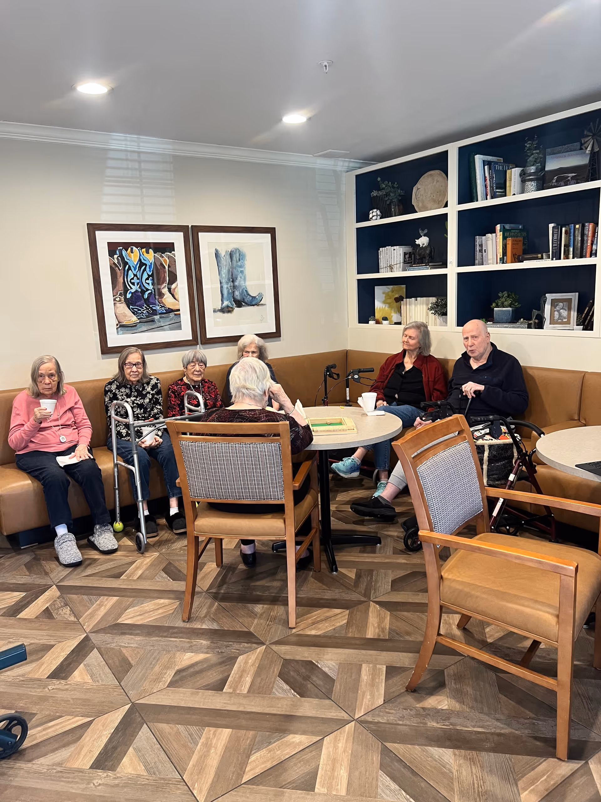A group of residents seated around a table in a communal living room with built-in bookshelves and framed artwork.