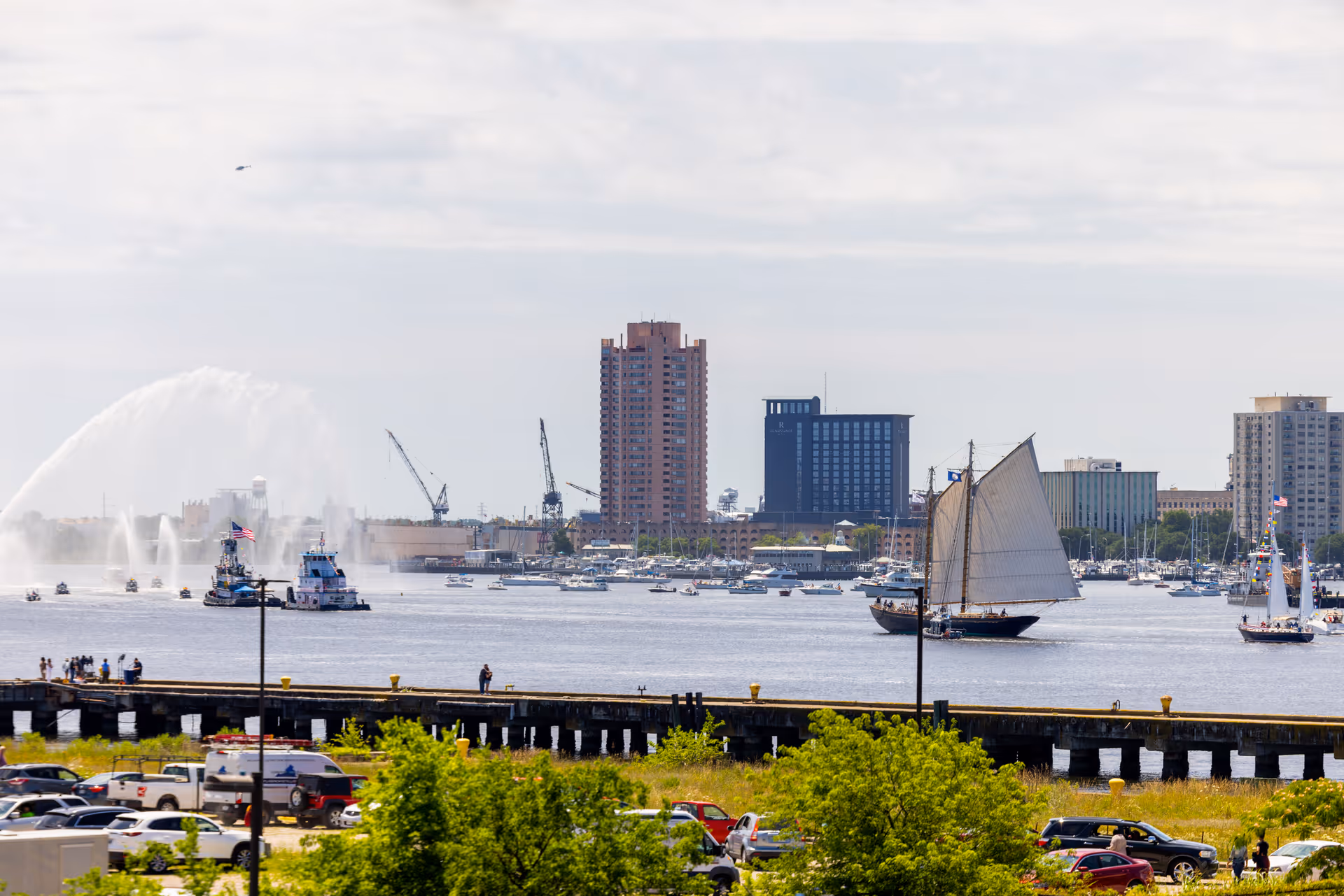 Harbor view with sailboats and a tugboat on the water, a pier and parked cars in the foreground, and city buildings along the opposite shore.