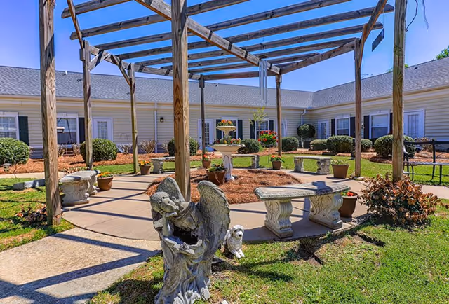 Outdoor courtyard area at Sumter Grove Senior Living featuring a wooden pergola, stone benches, potted plants, a decorative fountain, and angel and dog statues on a sunny day.