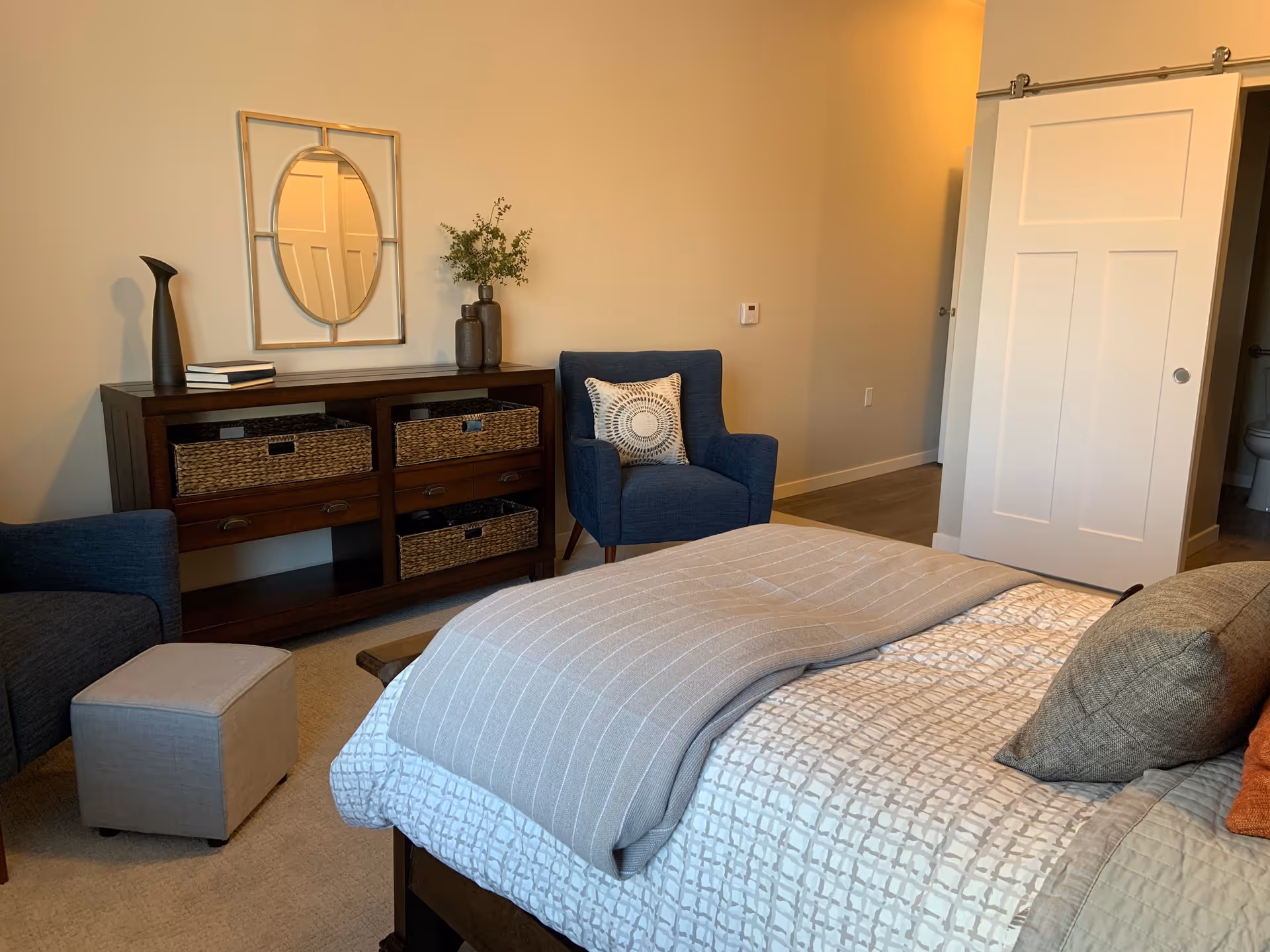 A cozy bedroom in a senior living facility featuring a neatly made bed with patterned bedding and multiple pillows. There are two blue armchairs, a small gray ottoman, and a dark wooden dresser with woven baskets and decorative items including a vase and books. A decorative mirror hangs on the wall above the dresser. A white sliding door is partially open, revealing a glimpse of a bathroom.