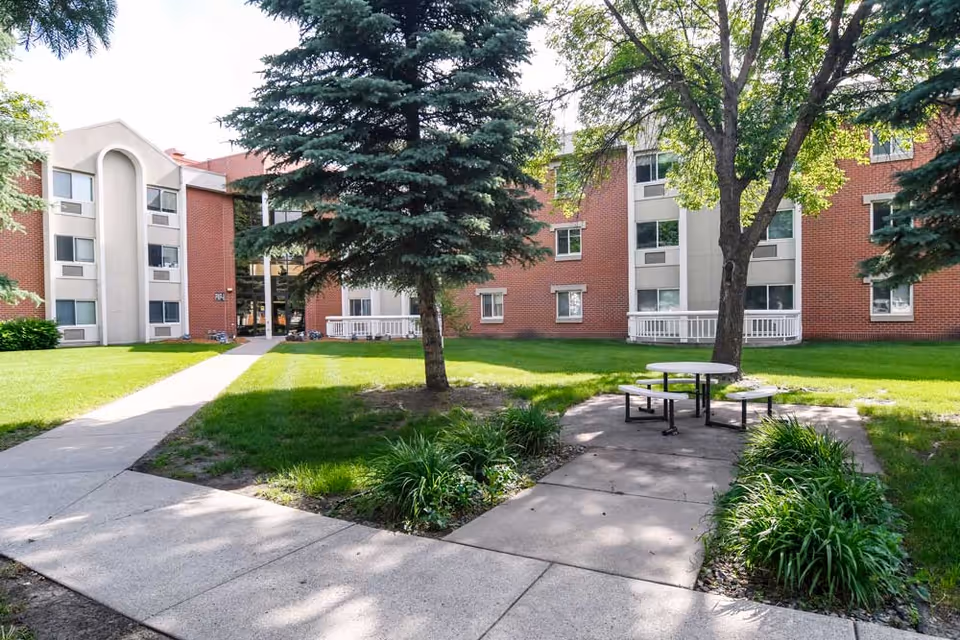 Outdoor courtyard area of a senior living facility with a concrete walkway, green grass, trees, and a round picnic table with attached benches. The background shows a red brick building with multiple windows and white trim.