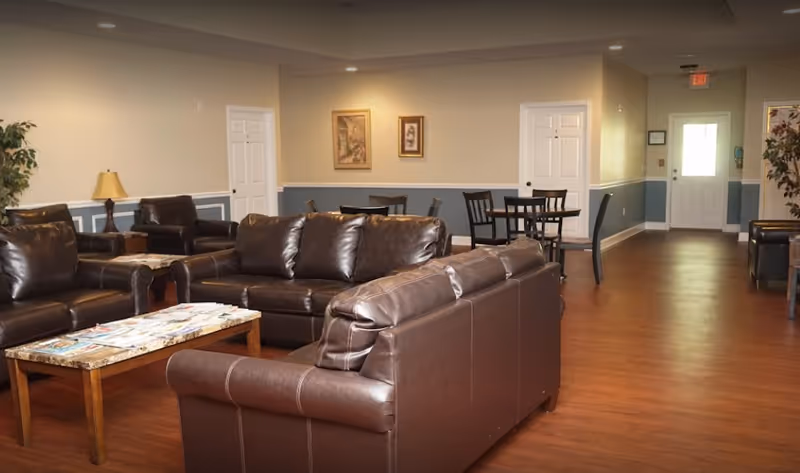 A spacious living room area with brown leather sofas and armchairs arranged around a wooden coffee table covered with magazines. The room has wooden flooring, beige walls with blue wainscoting, framed artwork on the walls, and a lamp on a side table. In the background, there are small dining tables with chairs and a door leading to another area.