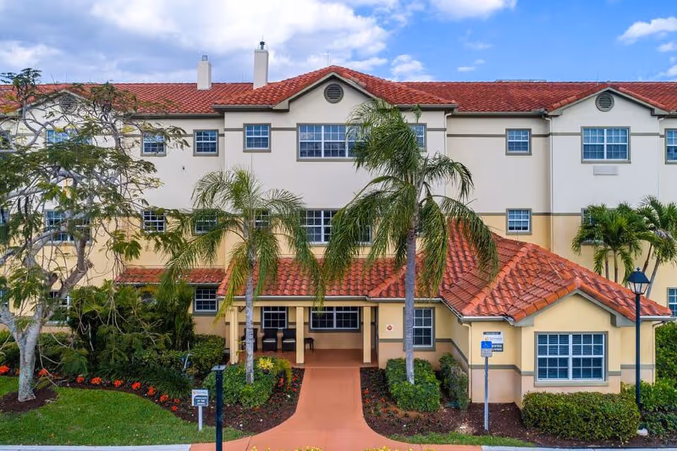 Front exterior of a three-story Mediterranean-style senior living building with a red tile roof, palm trees, and a landscaped entrance walkway.