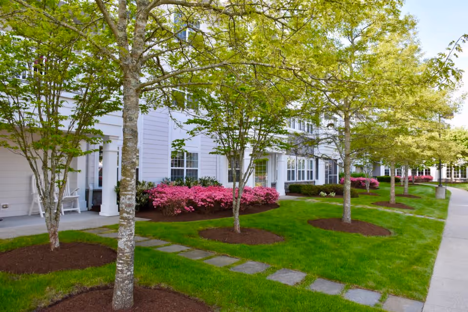 Exterior view of a senior living facility with a well-maintained lawn, several trees, pink flowering bushes, and a paved walkway leading to the building entrance. The building has white siding and multiple windows.