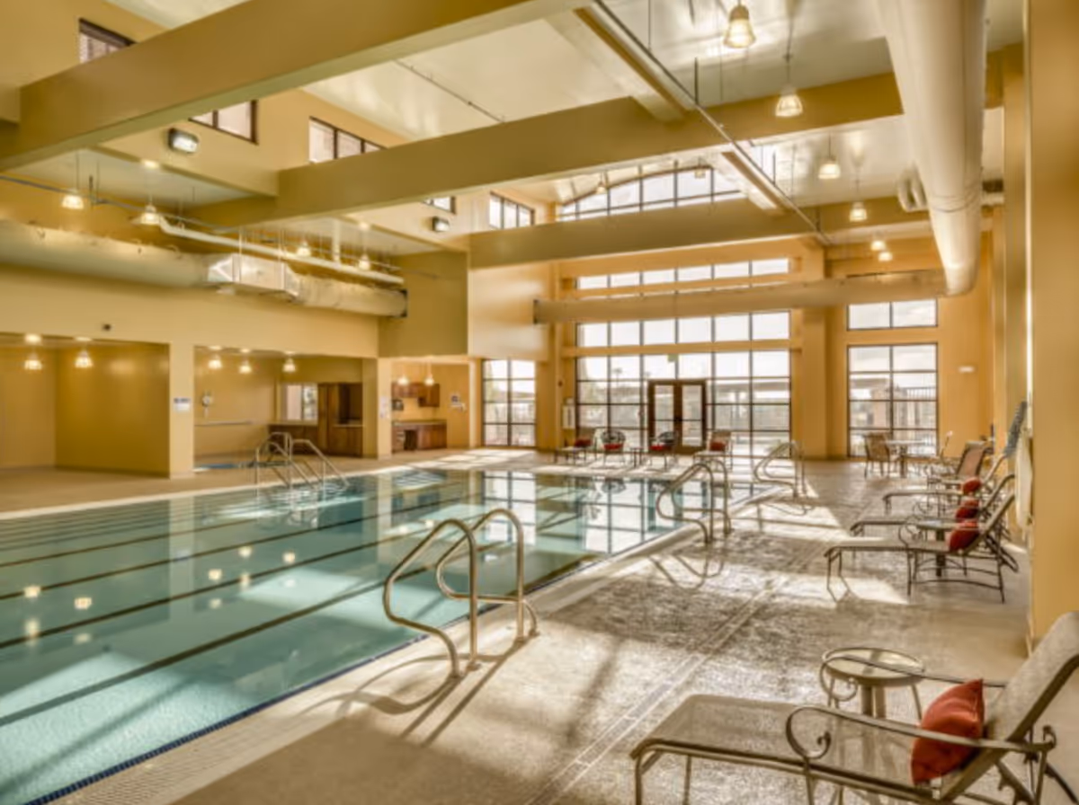 Indoor swimming pool area with multiple lanes, metal handrails, and seating along the right side with chairs and small tables. The space has high ceilings with exposed ducts and large windows letting in natural light.