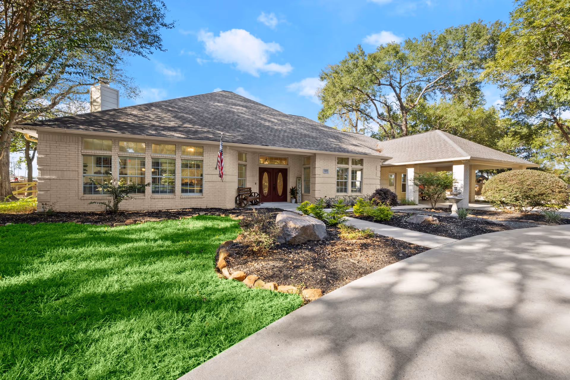 Exterior view of a single-story assisted living facility building with beige brick walls, large windows, and a covered entrance. The building is surrounded by green grass, landscaped garden beds with rocks and shrubs, and tall trees under a blue sky with scattered clouds.