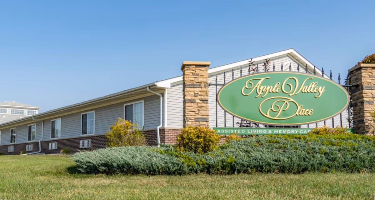 Exterior view of Apple Valley Place of Charles City, showing a single-story building with gray siding and brick accents, a well-maintained lawn, shrubs, and a large green sign with gold lettering that reads 'Apple Valley Place Assisted Living & Memory Care' under a clear blue sky.