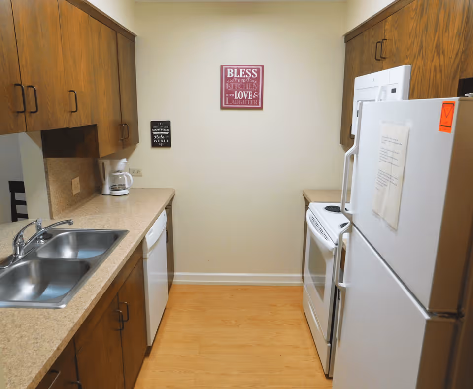 A small kitchen with wooden cabinets, a double sink, a white dishwasher, a white stove, a white refrigerator, and a coffee maker on the countertop. Two decorative signs are on the wall, one saying 'BLESS OUR KITCHEN WITH LOVE & LAUGHTER' and the other about coffee. The floor is light wood.