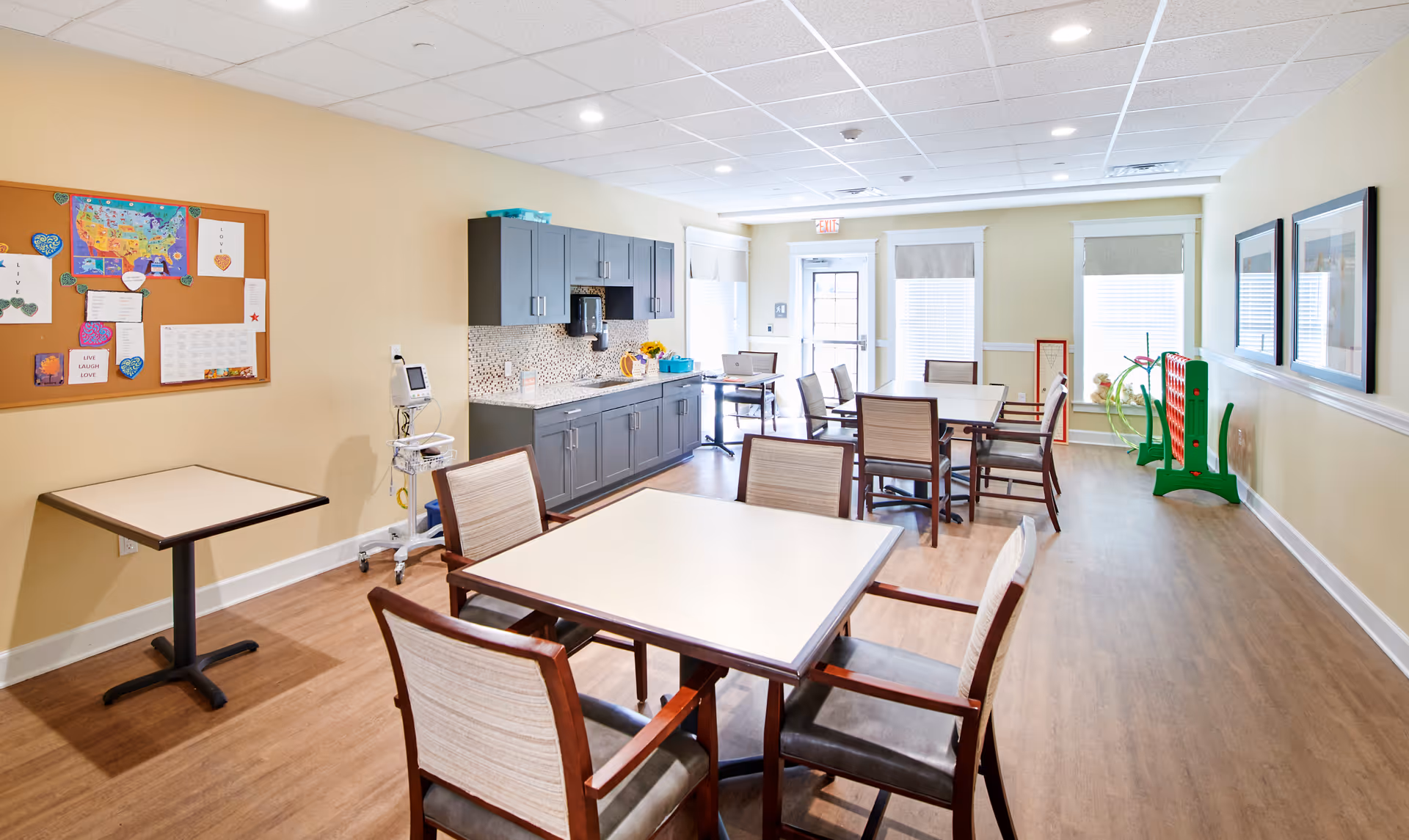 A bright and clean common room with several tables and chairs arranged for group seating. The room features a kitchenette area with gray cabinets, a countertop, and a coffee maker. A bulletin board with colorful papers and artwork is mounted on the left wall. Large windows with blinds allow natural light to fill the space. A large green Connect Four game is visible near the windows on the right side of the room.