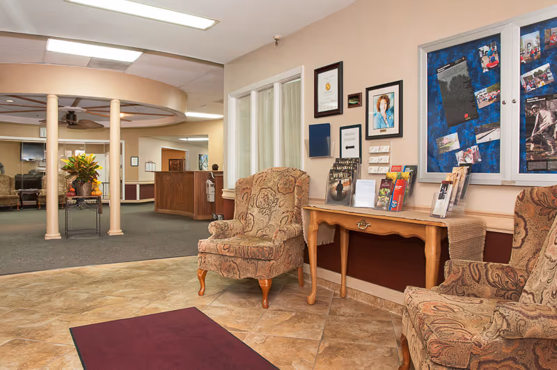 Interior view of a senior living facility lobby area with two patterned armchairs, a wooden table displaying brochures and pamphlets, framed certificates and photos on the wall, and a carpeted seating area with columns and additional chairs in the background.