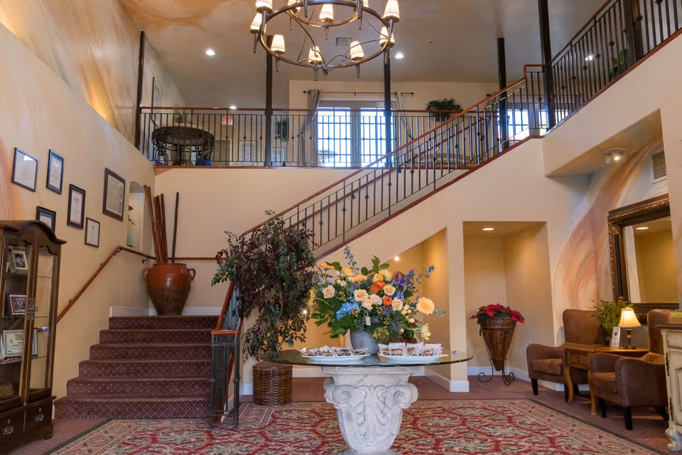 Interior view of a senior living facility lobby with a staircase leading to an upper floor. A round table with a large floral arrangement is centered on a patterned rug. There are two brown armchairs with a small table and lamp on the right side, and framed certificates and photos on the left wall. The area is warmly lit with a chandelier hanging from the ceiling.