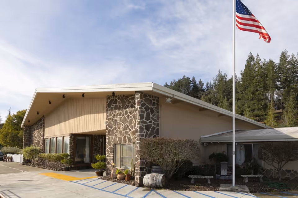 Front exterior of a single-story healthcare building with a stone facade, an American flag on a pole, and a paved, landscaped entrance.