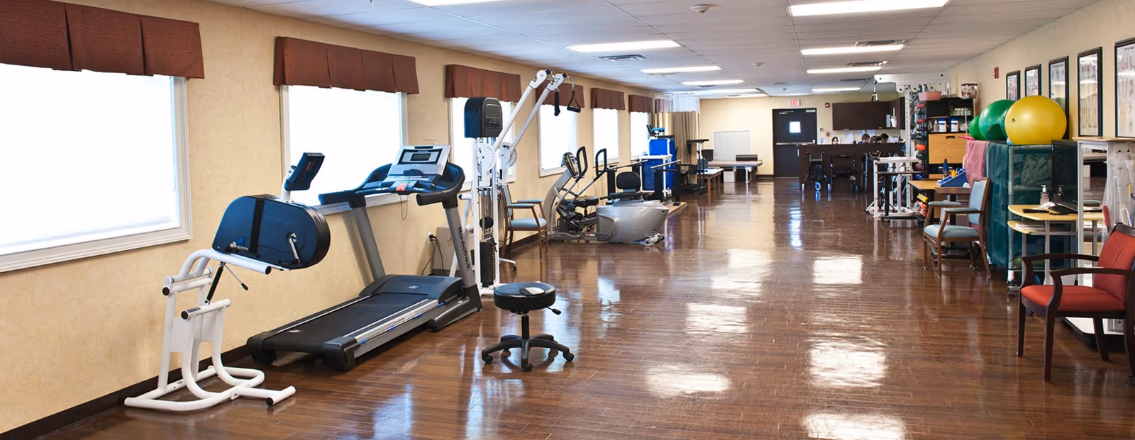 A spacious rehabilitation room with various exercise equipment including a treadmill, stationary bike, and other physical therapy machines. The room has large windows with brown valances, wooden flooring, and chairs along the walls. Exercise balls and therapy supplies are visible on the right side of the room.