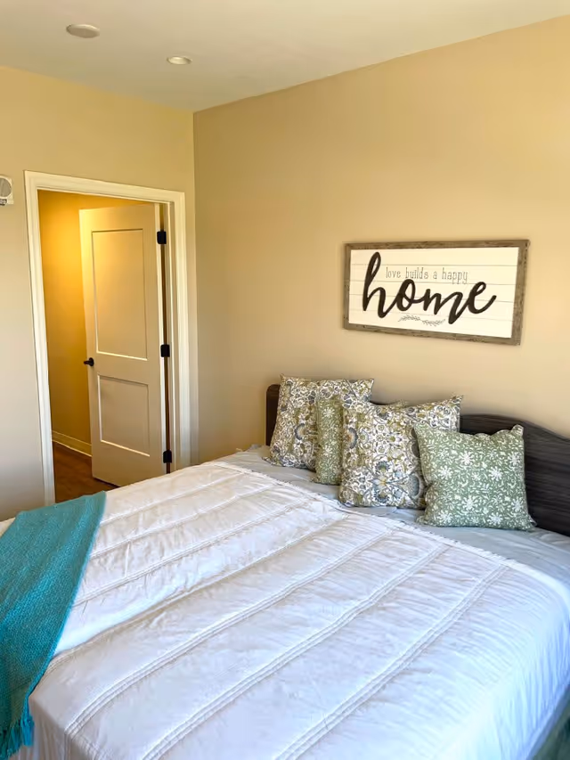 A bedroom with a neatly made bed featuring a white comforter, a teal throw blanket, and several decorative pillows in green and floral patterns. On the beige wall above the bed is a framed sign that reads 'love builds a happy home.' A white door is open, revealing a hallway with wooden flooring.