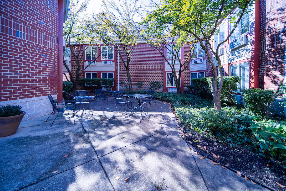 Outdoor courtyard area with metal tables and chairs surrounded by brick buildings and trees casting shadows on the concrete ground. There are green bushes and plants along the edges of the courtyard.