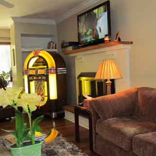 Cozy living room with a brown upholstered armchair, a small wooden side table with a lamp, a fireplace with a TV mounted above it, and a vintage jukebox in the corner. A green potted plant with white flowers is on a table in the foreground.