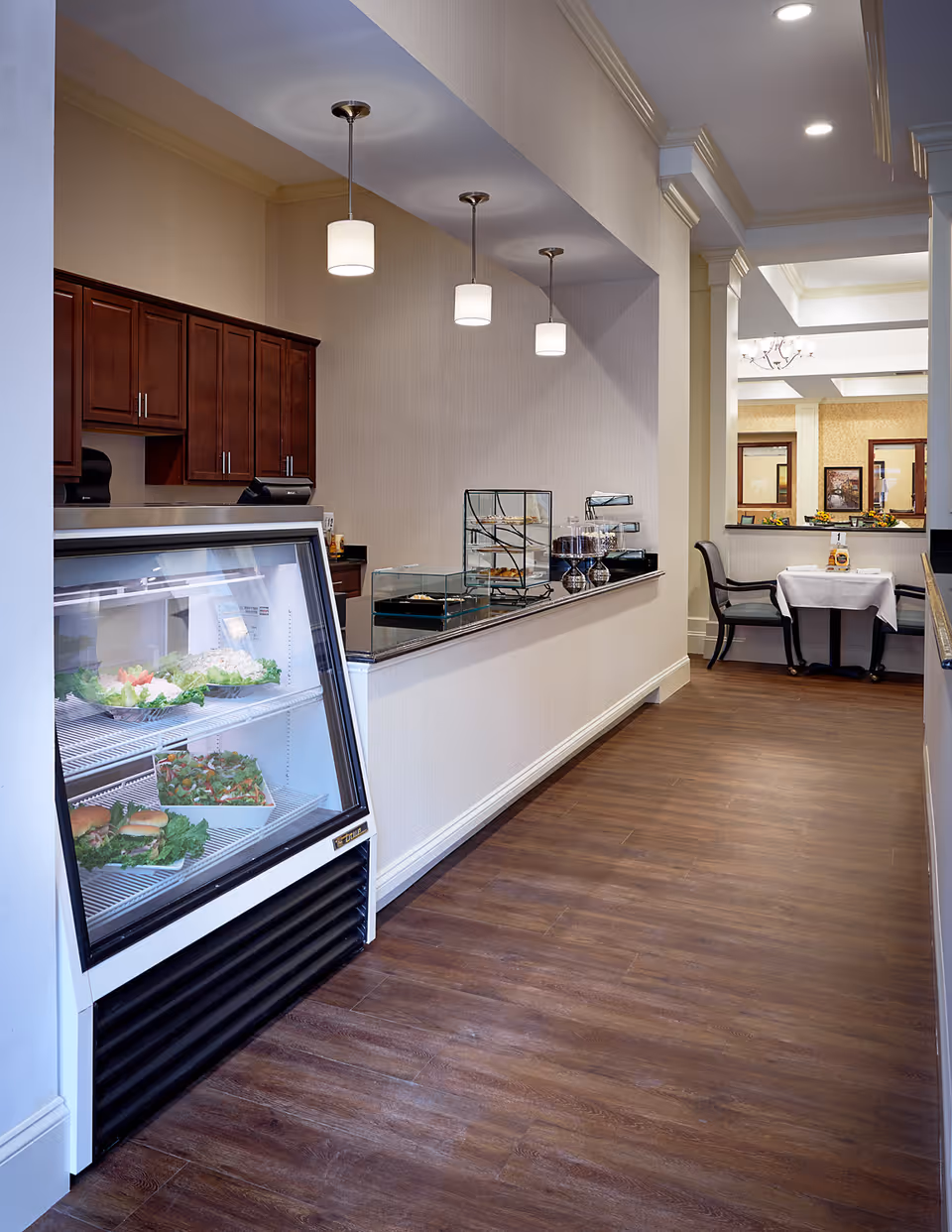 Interior view of a dining area in a senior living facility with a refrigerated display case containing sandwiches and salads on the left, a counter with glass food displays and three hanging pendant lights above it, and a small table with two chairs set for dining in the background.