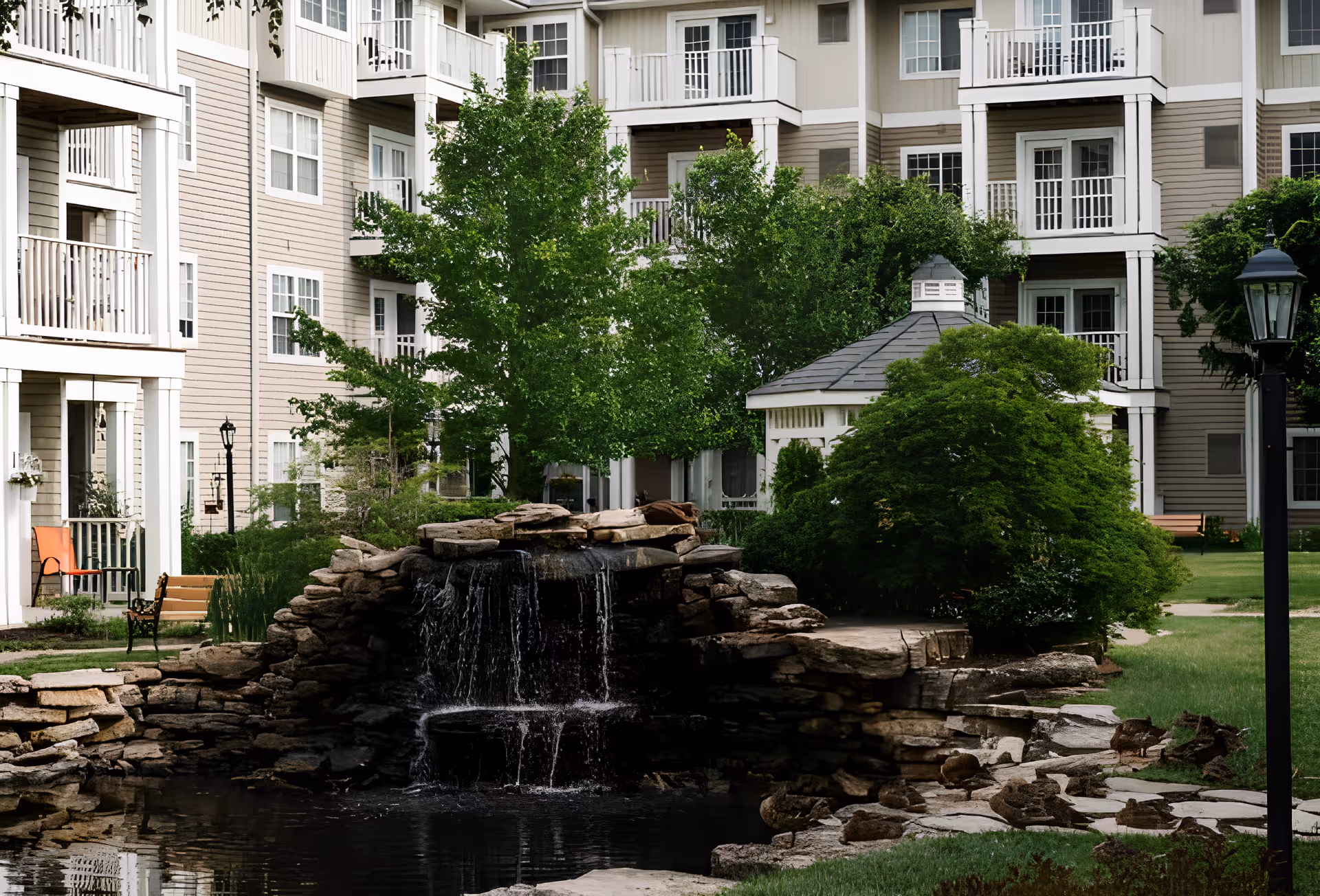 Courtyard of a senior living complex with a rock waterfall pond, gazebo, trees, and a multi-story building with balconies.