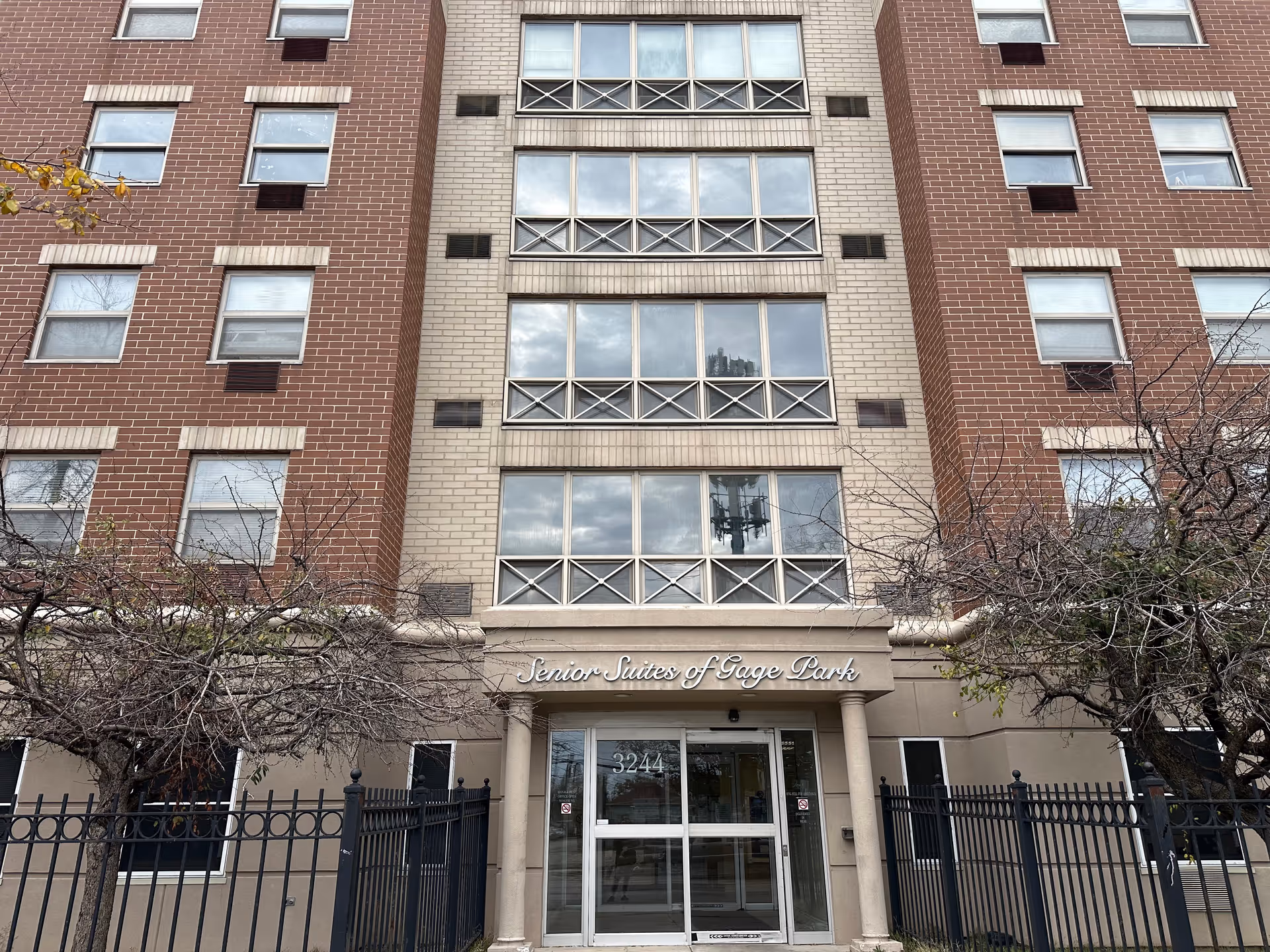 Front exterior view of a multi-story brick and beige building with the sign 'Senior Suites of Gage Park' above the entrance. The entrance has glass doors and is flanked by two columns. There are leafless trees and a black metal fence in front of the building.
