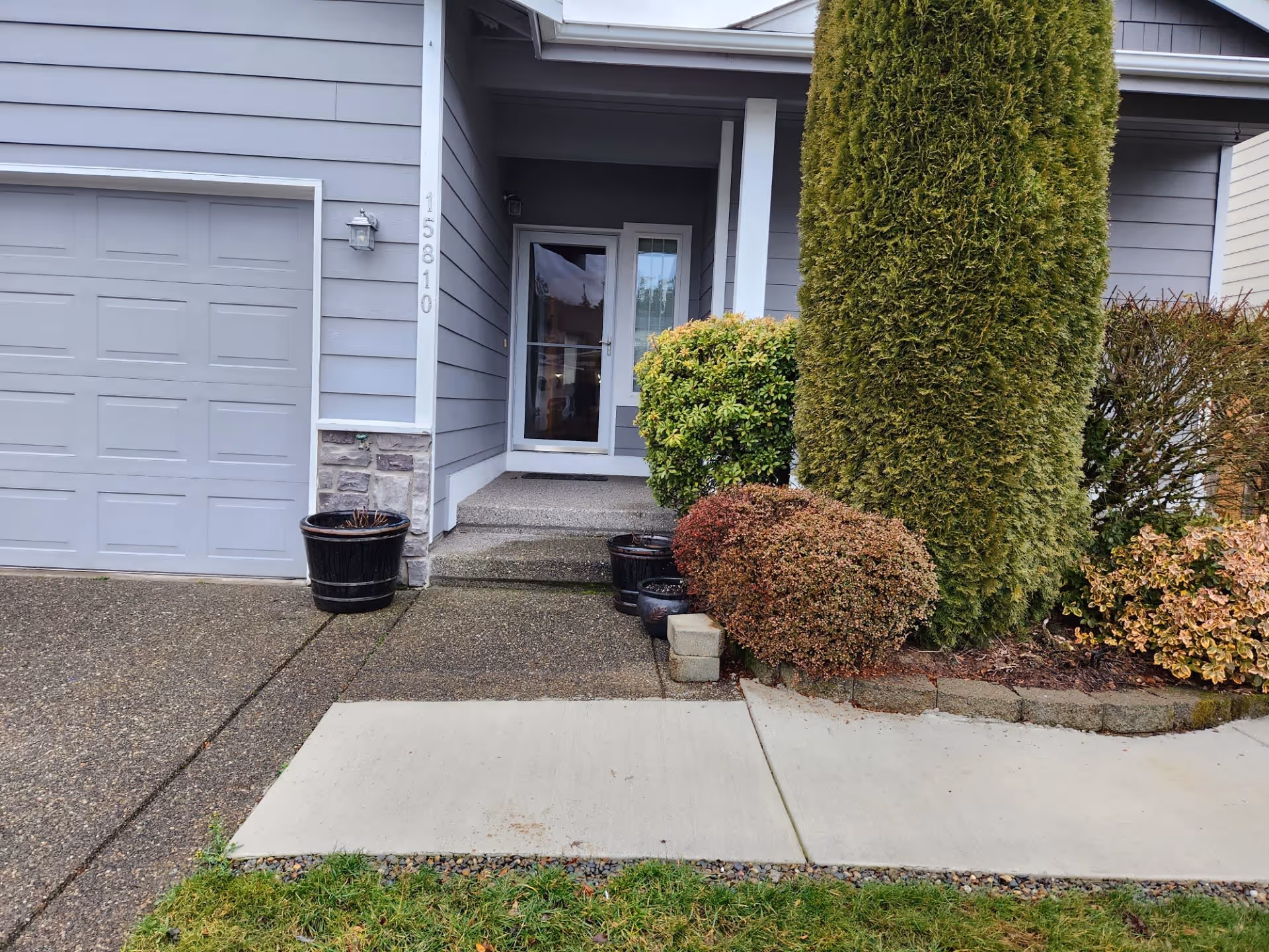 Front entrance of a residential building with a gray garage door on the left, a small porch with steps leading to a glass front door, and various shrubs and potted plants around the entrance.