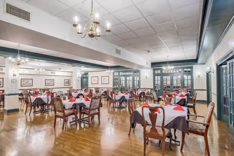 A spacious dining room with multiple tables covered in white and brown tablecloths, each set with red folded napkins and small floral centerpieces. The room features wooden chairs with patterned cushions, chandeliers hanging from the ceiling, and large windows with green trim along the walls.