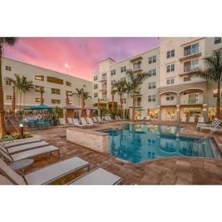 Outdoor swimming pool area at a senior living facility during sunset with lounge chairs arranged around the pool, palm trees, and a multi-story building in the background under a pink and purple sky.