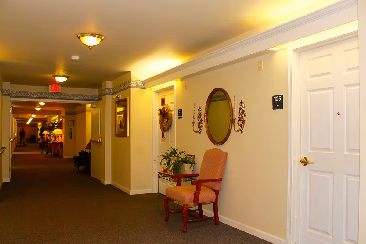 A well-lit hallway in a senior living facility with beige walls and carpeted floor. There are white doors with room numbers 125 and 127 visible, a decorative oval mirror with two wall sconces, a cushioned wooden chair, and a small table with plants. The hallway extends into the distance with additional lighting and furniture visible.