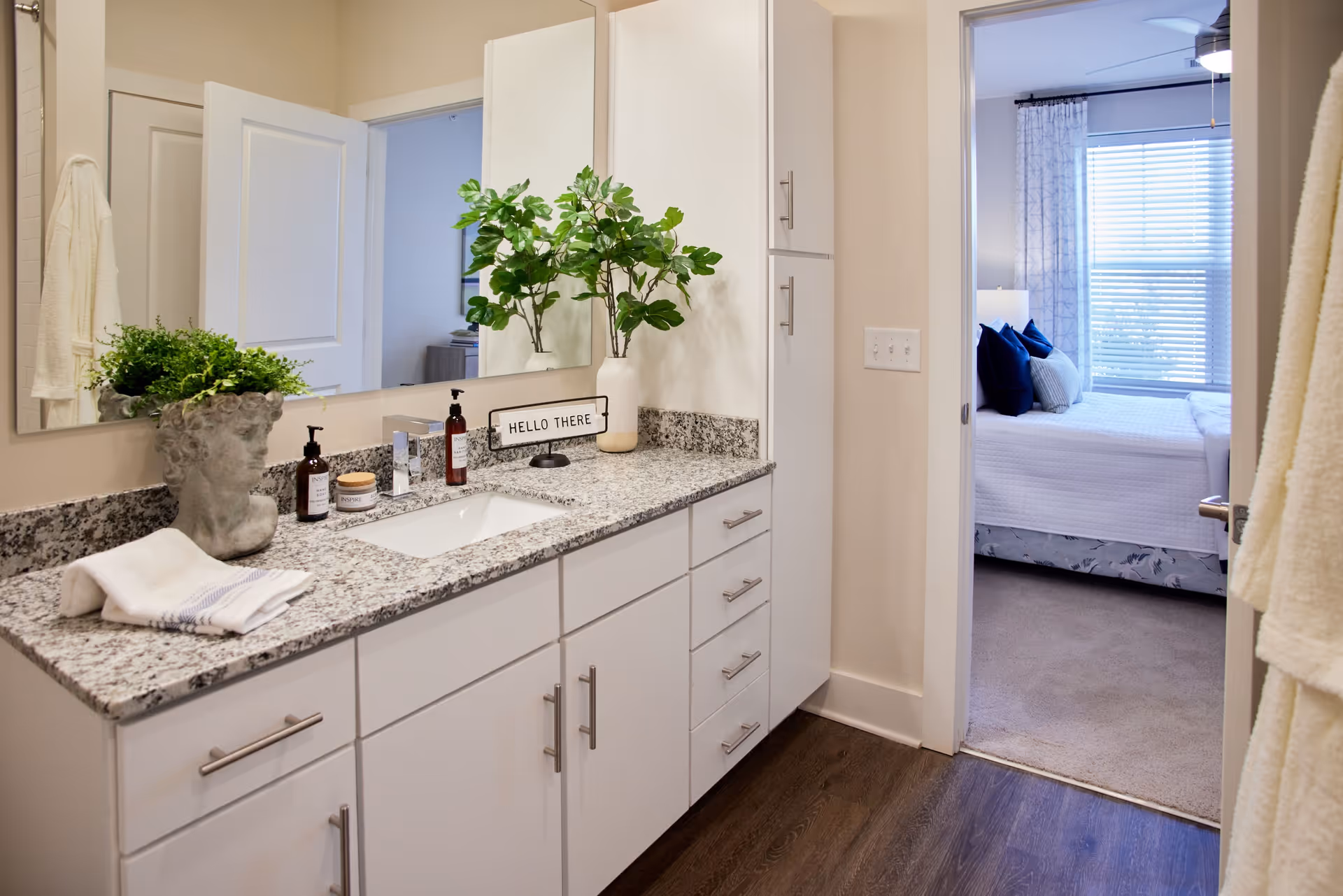A bathroom vanity with a granite countertop, white cabinets, a large mirror, and decorative items including a plant in a sculpted head planter, soap dispensers, and a sign that says 'HELLO THERE'. The bathroom opens into a bedroom with a bed, pillows, window blinds, and curtains.