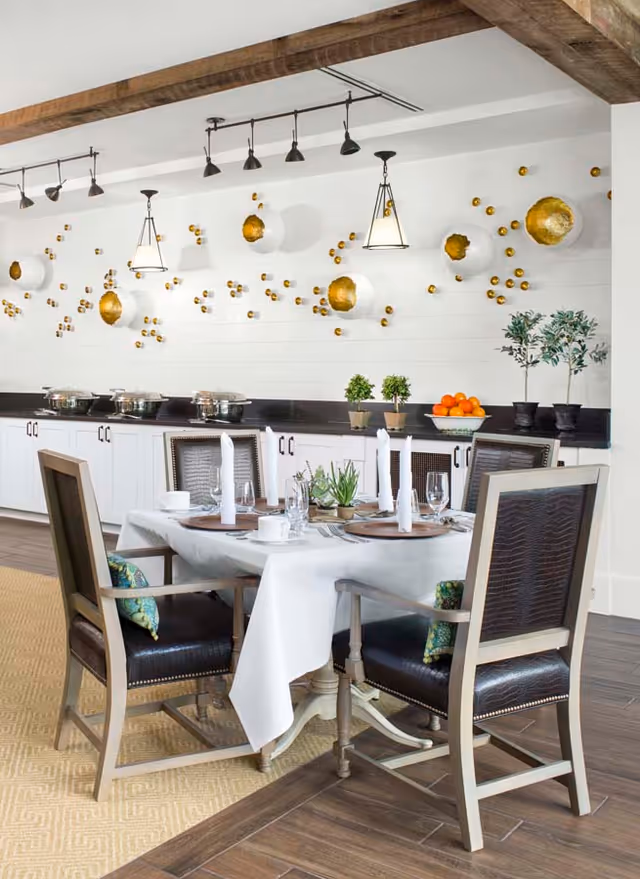 A dining area with a table set for four, featuring white tablecloth, napkins, glassware, and plates. The chairs have dark leather seats and backs with light wooden frames. Behind the table is a counter with white cabinets, decorative plants, a bowl of oranges, and several covered serving dishes. The wall above the counter is decorated with white and gold circular wall art and pendant lights hanging from the ceiling with exposed wooden beams.